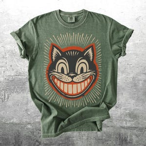 Op de afbeelding: Een saliegroen T-shirt met korte mouwen en een afbeelding van een zwarte kattenkop met een brede, tandige grijns. De kat is omlijnd in oranje en omringd door witte stralende lijnen. Het shirt heeft een ronde hals en een casual pasvorm.