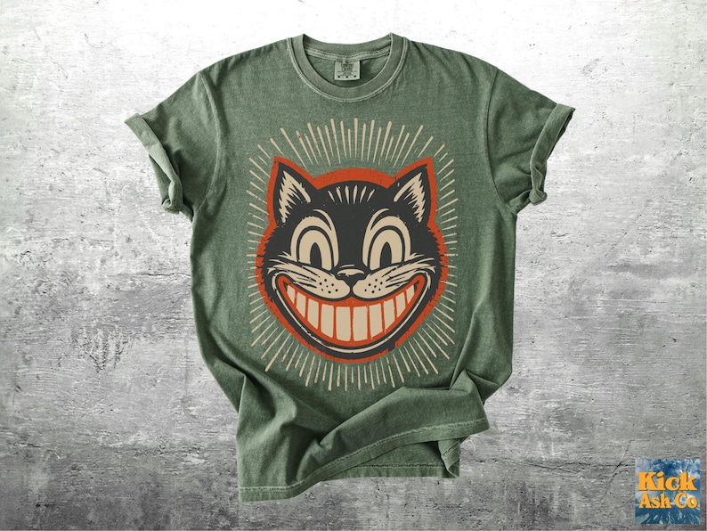 Op de afbeelding: Een saliegroen T-shirt met korte mouwen en een afbeelding van een zwarte kattenkop met een brede, tandige grijns. De kat is omlijnd in oranje en omringd door witte stralende lijnen. Het shirt heeft een ronde hals en een casual pasvorm.