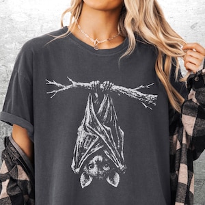 Op de afbeelding: Donkergrijs T-shirt met een witte illustratie van een vleermuis die aan een tak hangt. De vleermuis is gedetailleerd afgebeeld, met grote ogen en uitgespreide vleugels. Het shirt heeft korte mouwen en een relaxte pasvorm.