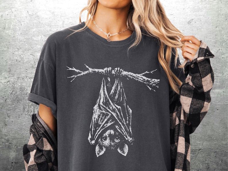 Op de afbeelding: Donkergrijs T-shirt met een witte illustratie van een vleermuis die aan een tak hangt. De vleermuis is gedetailleerd afgebeeld, met grote ogen en uitgespreide vleugels. Het shirt heeft korte mouwen en een relaxte pasvorm.