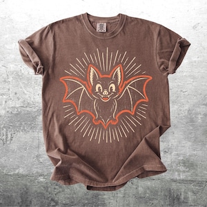 May include: A brown short-sleeve t-shirt featuring a central graphic of a smiling bat with outstretched wings. The bat is outlined in orange and surrounded by white radiating sunburst lines. The shirt has a crew neckline and rolled sleeves.