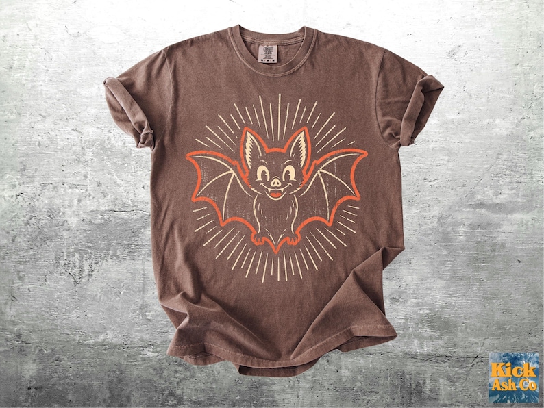 May include: A brown short-sleeve t-shirt featuring a central graphic of a smiling bat with outstretched wings. The bat is outlined in orange and surrounded by white radiating sunburst lines. The shirt has a crew neckline and rolled sleeves.