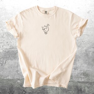 May include: A cream colored t-shirt with a black line drawing of a goose and the word "Silly" above it.