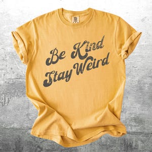 May include: Mustard yellow t-shirt with rolled-up sleeves. The shirt features the phrase "Be Kind Stay Weird" in a distressed black font. The shirt is made of a soft material and is likely a unisex fit.