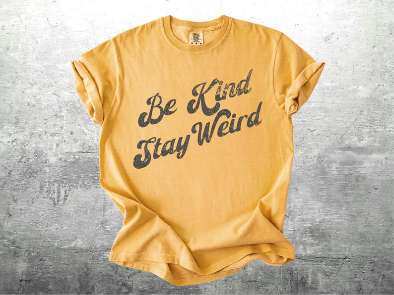 May include: Mustard yellow t-shirt with rolled-up sleeves. The shirt features the phrase "Be Kind Stay Weird" in a distressed black font. The shirt is made of a soft material and is likely a unisex fit.