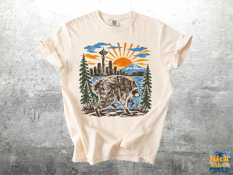 May include: A cream-coloured short-sleeve t-shirt featuring a graphic print of a raccoon walking in front of a landscape. The design includes the Seattle skyline with the Space Needle, a mountain, pine trees, and a bright orange sun in the sky.