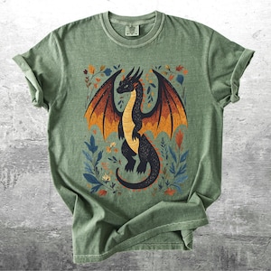 May include: A green t-shirt with a black dragon illustration surrounded by colorful flowers. The dragon has orange wings and is facing to the right.