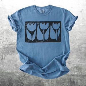 May include: A blue t-shirt with a black graphic of three stylized tulips in a rectangular frame. The shirt is made of soft material and has rolled-up sleeves. The design is centered on the front of the shirt.