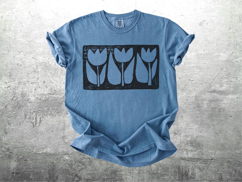 May include: A blue t-shirt with a black graphic of three stylized tulips in a rectangular frame. The shirt is made of soft material and has rolled-up sleeves. The design is centered on the front of the shirt.