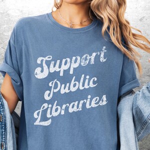 May include: A blue t-shirt with the text "Support Public Libraries" in a white, vintage-style font. The shirt has rolled-up sleeves and is paired with a denim jacket.