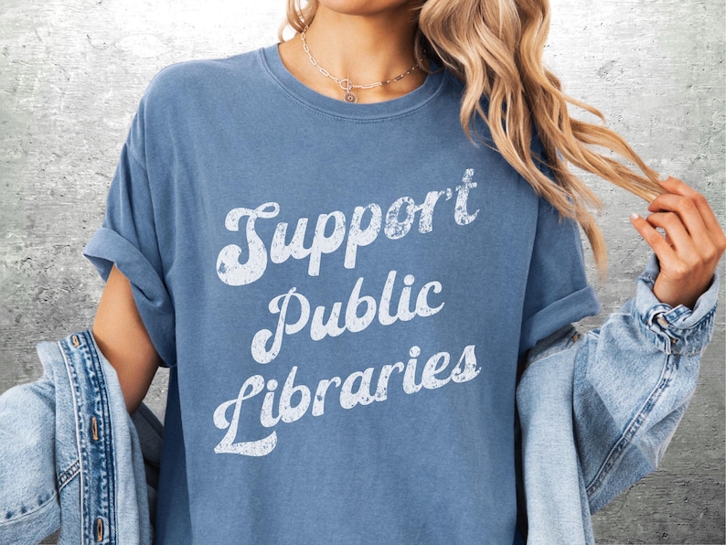 May include: A blue t-shirt with the text "Support Public Libraries" in a white, vintage-style font. The shirt has rolled-up sleeves and is paired with a denim jacket.