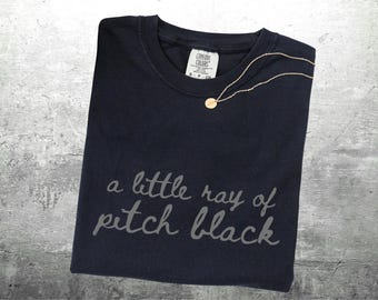 May include: A navy blue t-shirt with the phrase "a little ray of pitch black" in light gray script. A gold-colored necklace with a small pendant rests on the shirt. The shirt has a Comfort Colors tag.