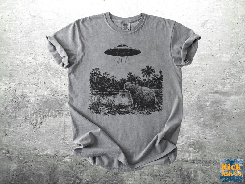 May include: A gray t-shirt featuring a black and white graphic of a capybara standing near a body of water with a UFO hovering above. The shirt has rolled-up sleeves and a crew neck. The text "Kick Ash Co" is in the bottom right corner.