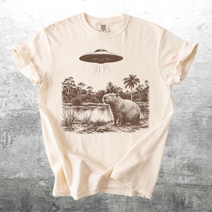 May include: Cream-colored t-shirt featuring a sepia-toned graphic of a capybara standing near a body of water under a hovering UFO. Palm trees and other foliage are in the background. The shirt has rolled-up sleeves.