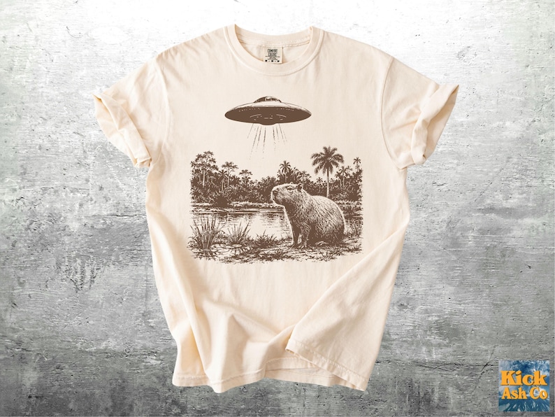 May include: Cream-colored t-shirt featuring a sepia-toned graphic of a capybara standing near a body of water under a hovering UFO. Palm trees and other foliage are in the background. The shirt has rolled-up sleeves.