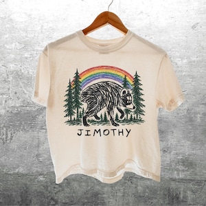 May include: A cream-colored short-sleeve t-shirt featuring a graphic of a black-and-white raccoon walking through a forest of green pine trees under a colorful rainbow. The text JIMOTHY is printed in black capital letters below the illustration.