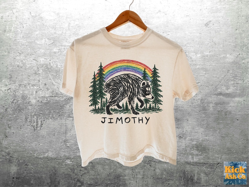 May include: A cream-colored short-sleeve t-shirt featuring a graphic of a black-and-white raccoon walking through a forest of green pine trees under a colorful rainbow. The text JIMOTHY is printed in black capital letters below the illustration.