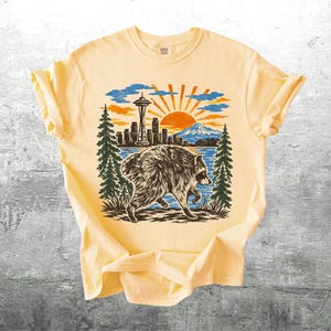 May include: A pale yellow short-sleeve t-shirt featuring a graphic print of a raccoon walking in front of a Seattle skyline, including the Space Needle, a mountain, and a sunburst. The design uses black, orange, and blue ink colours.