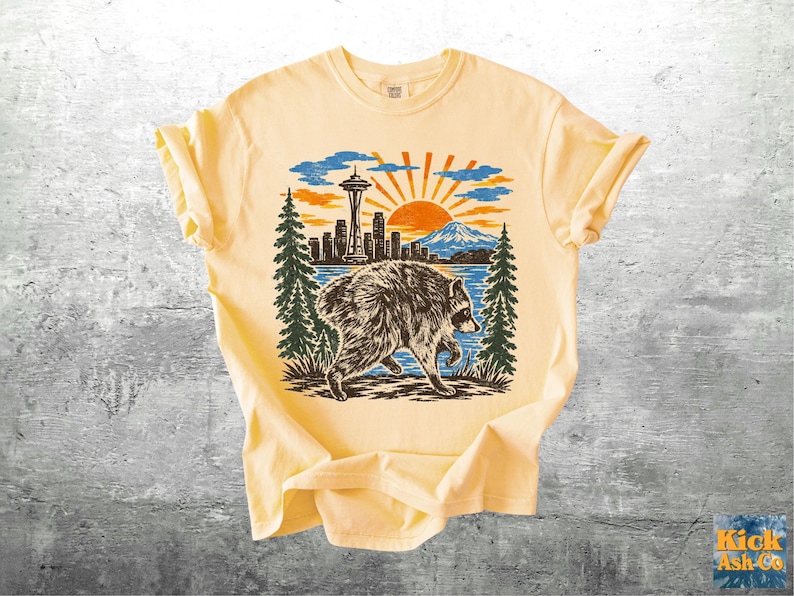 May include: A pale yellow short-sleeve t-shirt featuring a graphic print of a raccoon walking in front of a Seattle skyline, including the Space Needle, a mountain, and a sunburst. The design uses black, orange, and blue ink colours.