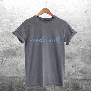 May include: A charcoal grey t-shirt with the text "needs salt" written in blue.