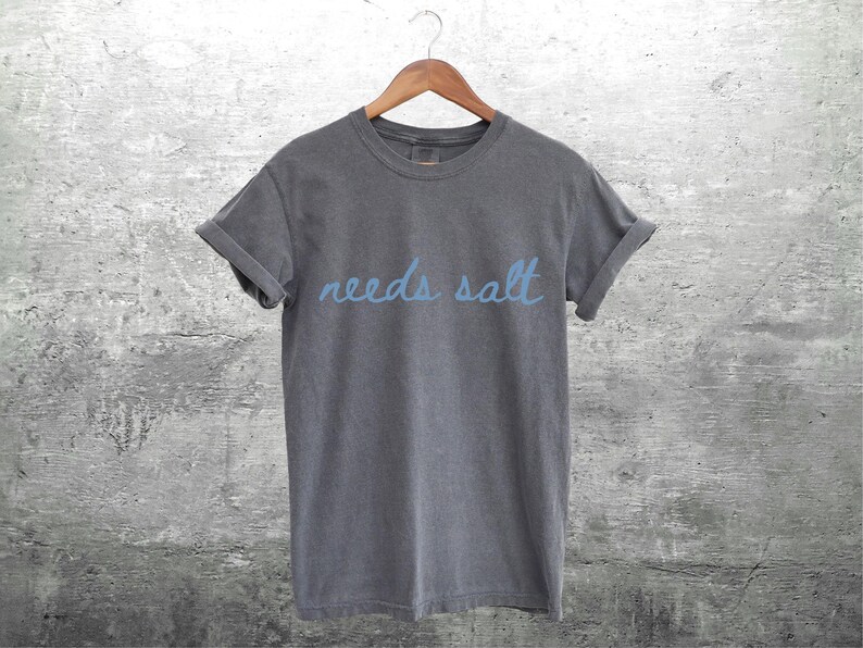 May include: A charcoal grey t-shirt with the text "needs salt" written in blue.