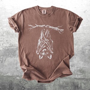 Op de afbeelding: Bruin t-shirt met een witte illustratie van een vleermuis die ondersteboven aan een tak hangt. Het t-shirt heeft een ronde hals en korte mouwen. De stof heeft een licht vervaagde uitstraling.
