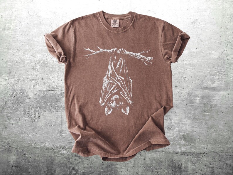 Op de afbeelding: Bruin t-shirt met een witte illustratie van een vleermuis die ondersteboven aan een tak hangt. Het t-shirt heeft een ronde hals en korte mouwen. De stof heeft een licht vervaagde uitstraling.