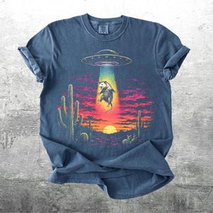 May include: A blue short-sleeve t-shirt featuring a graphic of an opossum being abducted by a UFO in a desert landscape at sunset. The scene includes cacti, a bright sun, and a colorful beam of light. The text Kick Ash Co is in the corner.