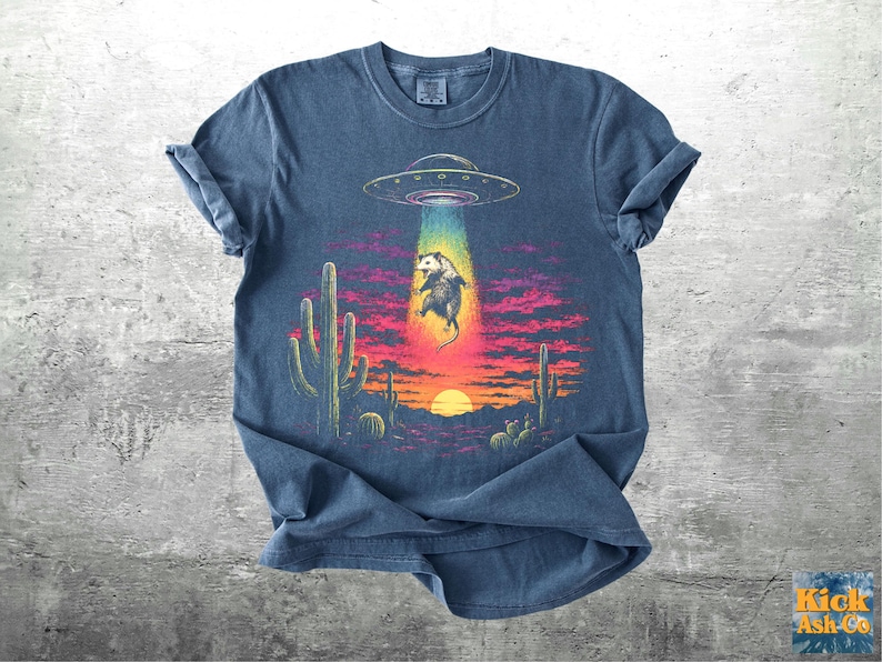 May include: A blue short-sleeve t-shirt featuring a graphic of an opossum being abducted by a UFO in a desert landscape at sunset. The scene includes cacti, a bright sun, and a colorful beam of light. The text Kick Ash Co is in the corner.