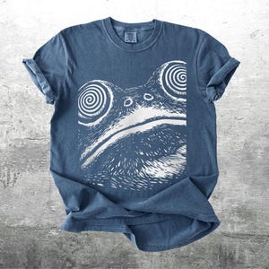 May include: A blue t-shirt featuring a white graphic of a frog's face with spiral eyes. The shirt has rolled-up sleeves and a crew neck. The frog design is centered on the front of the shirt, creating a unique and eye-catching design.