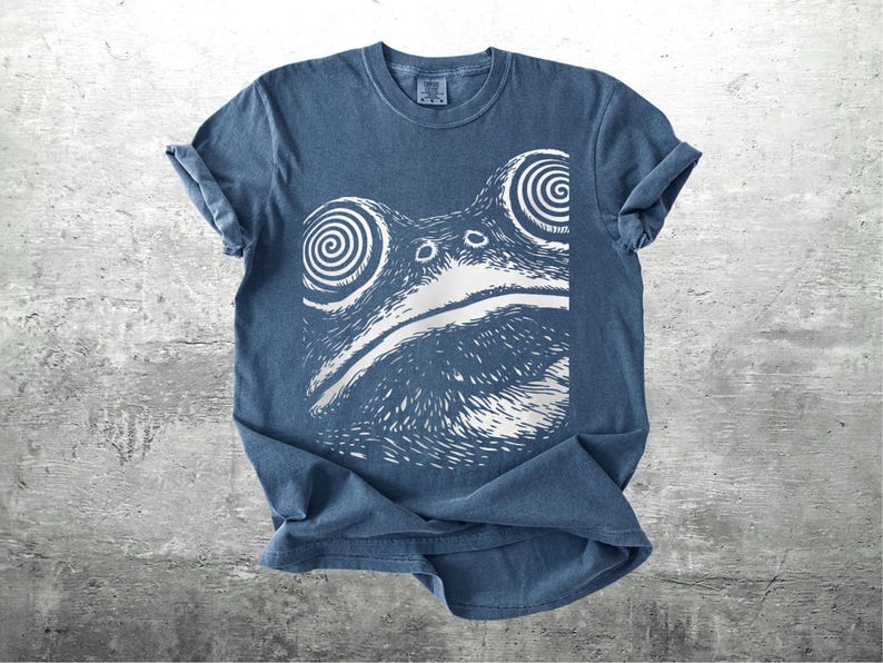 May include: A blue t-shirt featuring a white graphic of a frog's face with spiral eyes. The shirt has rolled-up sleeves and a crew neck. The frog design is centered on the front of the shirt, creating a unique and eye-catching design.