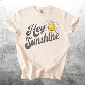 May include: Cream-colored t-shirt with the phrase "Hey Sunshine" in distressed gray lettering. A yellow sun graphic with radiating lines is to the right of the text. The shirt has short sleeves and a crew neck.