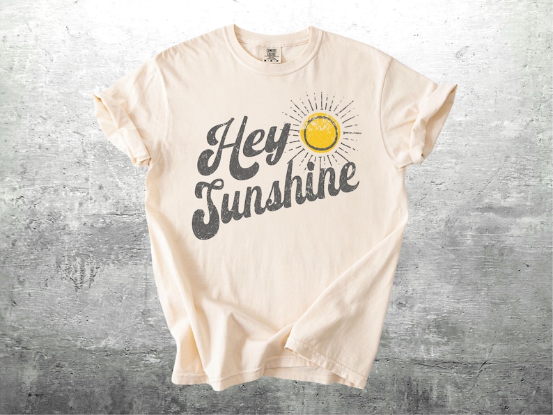 May include: Cream-colored t-shirt with the phrase "Hey Sunshine" in distressed gray lettering. A yellow sun graphic with radiating lines is to the right of the text. The shirt has short sleeves and a crew neck.