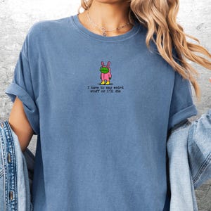 May include: Blue t-shirt with a small graphic of a frog in a pink bunny costume and the text "I have to say weird stuff or I'll die". The shirt has short sleeves and a crew neck.