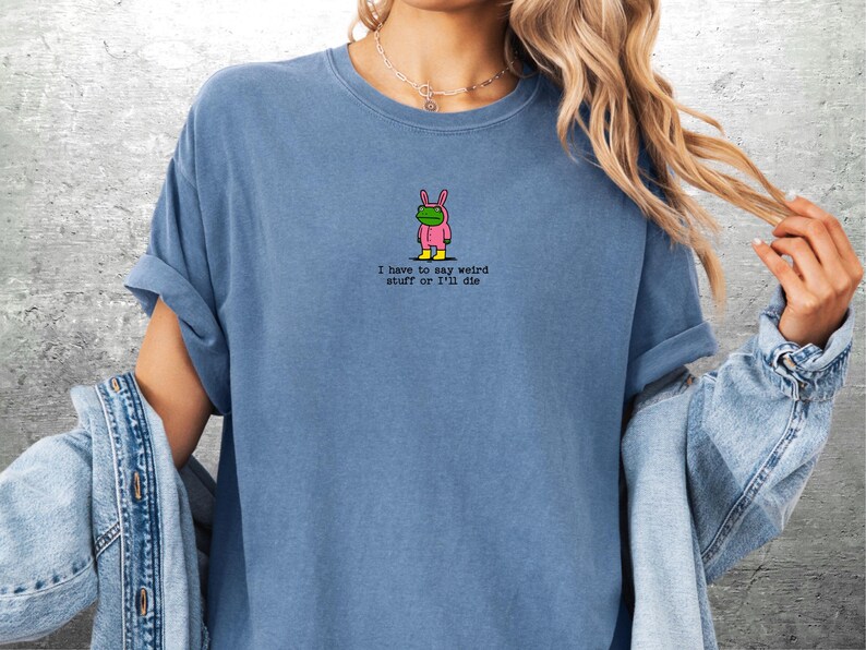 May include: Blue t-shirt with a small graphic of a frog in a pink bunny costume and the text "I have to say weird stuff or I'll die". The shirt has short sleeves and a crew neck.