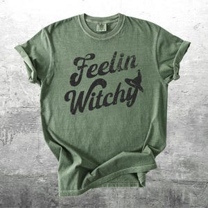 K&ouml;nnte beinhalten: Olivgr&uuml;nes T-Shirt mit hochgekrempelten &Auml;rmeln. Das Shirt zeigt den Schriftzug "Feelin Witchy" in einer verblassten schwarzen Schrift, mit einer Hexenhut-Grafik. Das T-Shirt ist aus weichem Material.