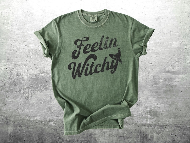 K&ouml;nnte beinhalten: Olivgr&uuml;nes T-Shirt mit hochgekrempelten &Auml;rmeln. Das Shirt zeigt den Schriftzug "Feelin Witchy" in einer verblassten schwarzen Schrift, mit einer Hexenhut-Grafik. Das T-Shirt ist aus weichem Material.