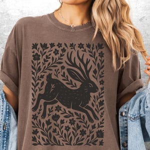 May include: Brown t-shirt featuring a black illustration of a running hare with antlers, surrounded by floral and leaf elements. The design is centered on the shirt. The shirt has rolled-up sleeves and a relaxed fit.