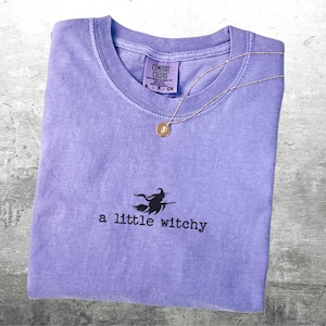 May include: A lavender Comfort Colors t-shirt with the phrase "a little witchy" and a witch graphic. A gold-colored necklace with a pendant rests on the shirt. The shirt is folded and displayed on a gray surface.