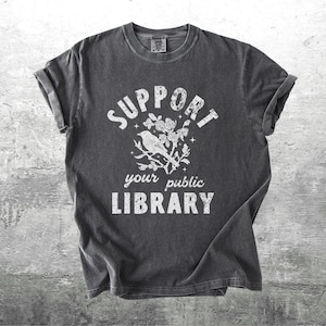 May include: Dark gray t-shirt with rolled sleeves. The shirt features the text "SUPPORT your public LIBRARY" in white, along with a graphic of a bird perched on a floral branch. The shirt is displayed on a gray concrete background.