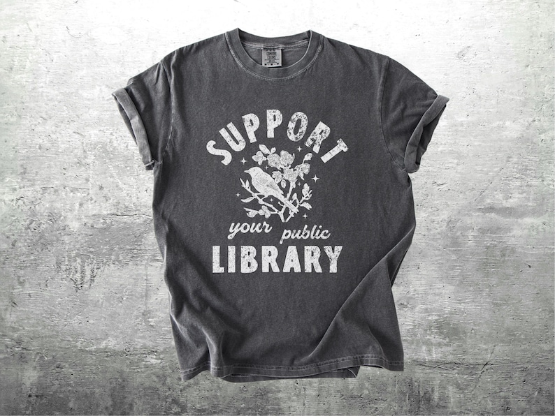 May include: Dark gray t-shirt with rolled sleeves. The shirt features the text "SUPPORT your public LIBRARY" in white, along with a graphic of a bird perched on a floral branch. The shirt is displayed on a gray concrete background.