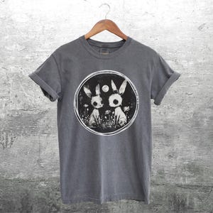 Op de afbeelding: Een donkergrijs T-shirt met een ronde afbeelding van twee witte konijnen met zwarte ogen en een volle maan op de achtergrond. De konijnen staan in een veld met bloemen.