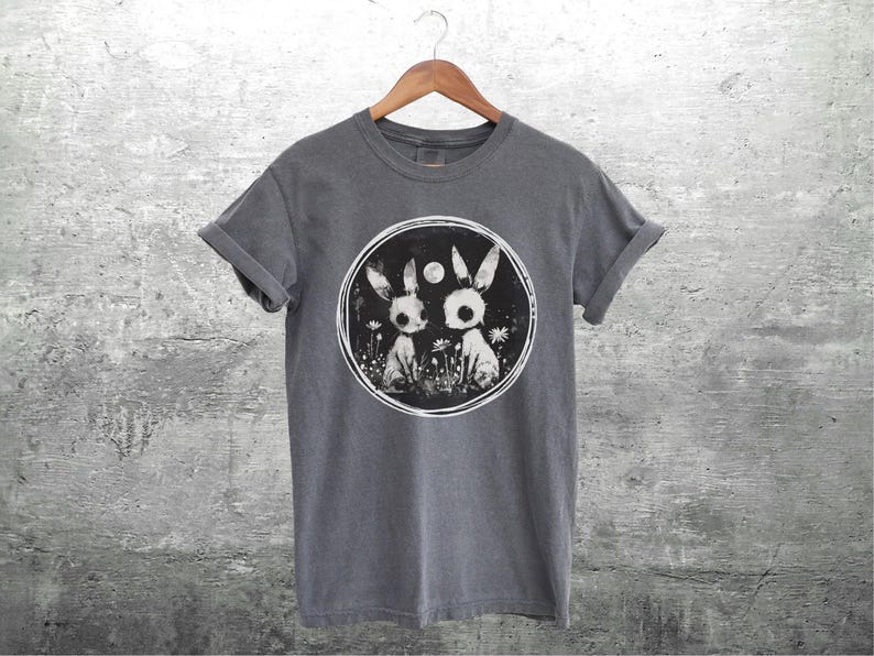 Op de afbeelding: Een donkergrijs T-shirt met een ronde afbeelding van twee witte konijnen met zwarte ogen en een volle maan op de achtergrond. De konijnen staan in een veld met bloemen.
