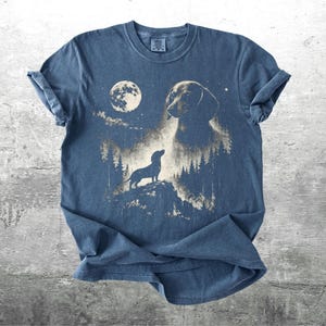 May include: Blue t-shirt featuring a dachshund dog design. The graphic includes a full moon, a silhouette of a dog on a hill, and a larger dog head. The design is in shades of tan and white.