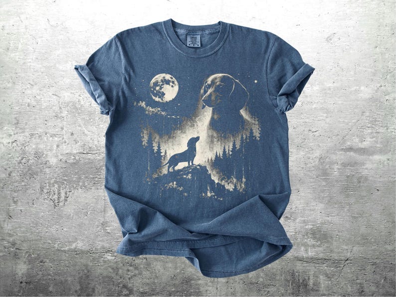 May include: Blue t-shirt featuring a dachshund dog design. The graphic includes a full moon, a silhouette of a dog on a hill, and a larger dog head. The design is in shades of tan and white.