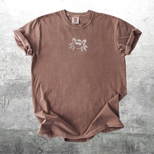 May include: A brown t-shirt with a small, white graphic on the chest. The shirt has short sleeves and a crew neck. The fabric appears soft and slightly faded, giving it a vintage look.