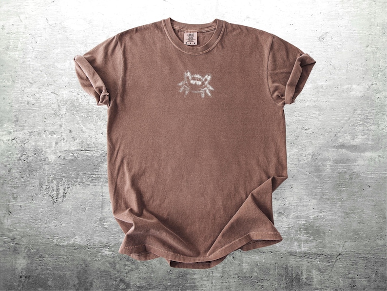 May include: A brown t-shirt with a small, white graphic on the chest. The shirt has short sleeves and a crew neck. The fabric appears soft and slightly faded, giving it a vintage look.