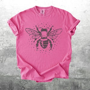 May include: A pink t-shirt featuring a large, detailed illustration of a bee with outstretched wings and a starburst design. The bee is dark gray, and the shirt has rolled-up sleeves.