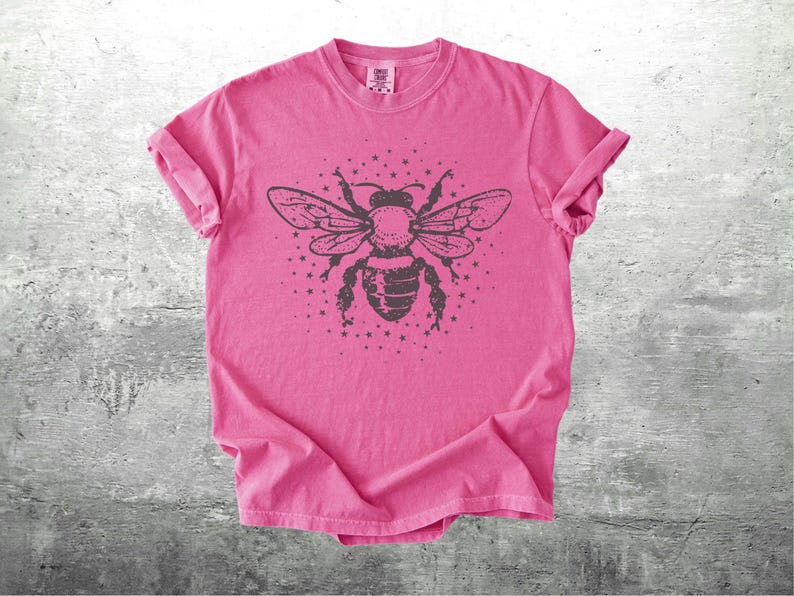 May include: A pink t-shirt featuring a large, detailed illustration of a bee with outstretched wings and a starburst design. The bee is dark gray, and the shirt has rolled-up sleeves.