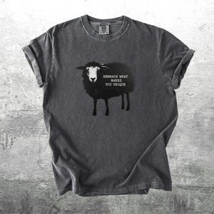 Puede incluir: Camiseta gris oscuro con una ilustraci&oacute;n de oveja negra y el texto "EMBRACE WHAT MAKES YOU UNIQUE." La camiseta tiene mangas enrolladas y cuello redondo. La camiseta est&aacute; hecha de un material suave y c&oacute;modo.